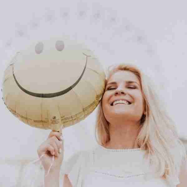 woman smiling holding a smiley face balloon showing why it pays to be positive